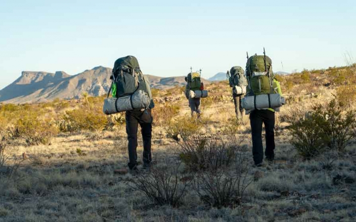 a group of gap year students carrying backpacks hike across a desert landscape toward a mountain in the distance  
